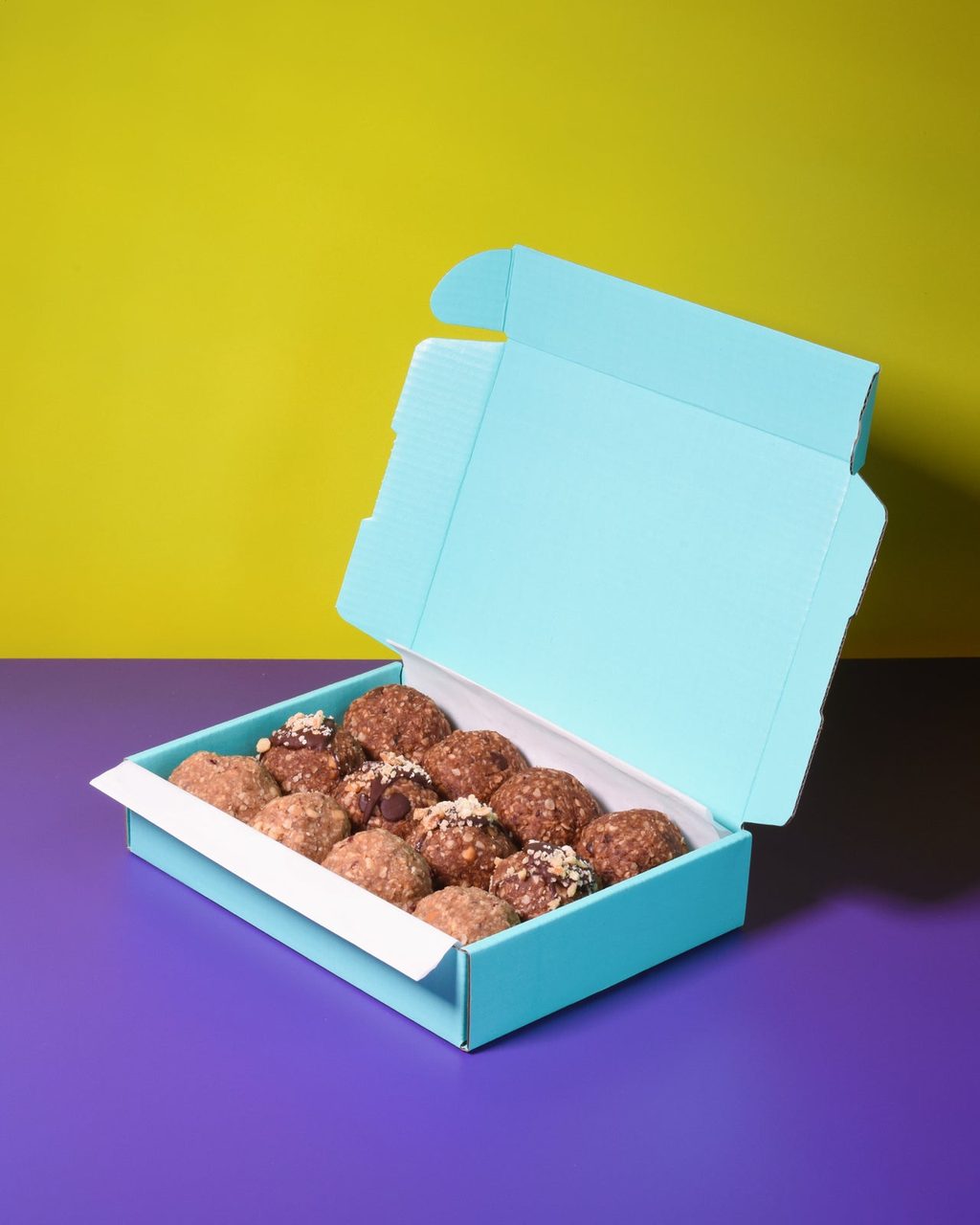 An open turquoise postal box of twelve raw energy balls on a purple surface in front of a yellow wall.