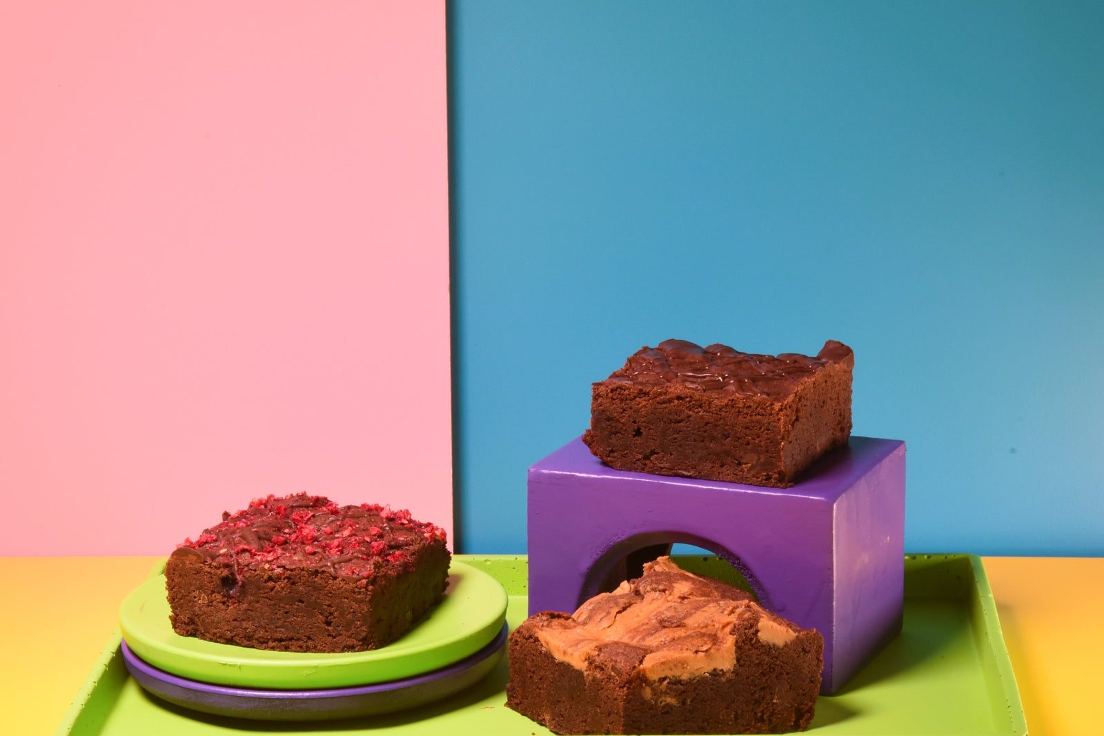 Three brownies, raspberry, chocolate and peanut butter swirl, on a green tray in front of pink and blue panels.